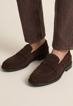 Brune ruskind herre loafers båret med matchende brune bukser på en lys beige baggrund.