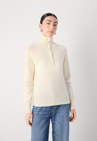 Pull en tricot couleur crème avec col montant, quatre grands boutons et poignets côtelés. Texture lisse avec des détails subtils sur les manches.