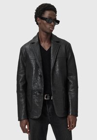 Schwarze, strukturiertes Lederjacke mit maßgeschneidertem Design, ausgestattet mit einem Reverskragen, zwei Knöpfen und zwei vorderen Taschen. Getragen über einem schwarzen Pullover.