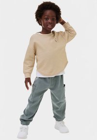 Beige sweatshirt med rund halsudskæring og lille logo, kombineret med lysegrønne joggers med sidelommer. Hvide sneakers fuldender outfittet.