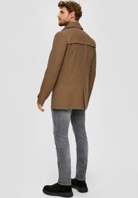 Braune Jacke mit strukturiertem Design und Rückeneinsatz, kombiniert mit grauen Jeans und schwarzen Stiefeln. Glatte Textur und taillierte Passform.