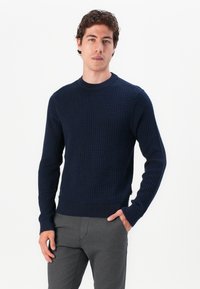 Dunkelblauer Strickpullover mit einem strukturierten Muster, Rundhalsausschnitt und gerippten Bündchen und Saum. Kombiniert mit grauen Hosen, die Hände in den Taschen.