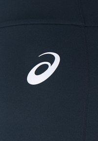 Vit Asics-logotyp på mörk marinblå atletisk tyg med synlig sömsöm.