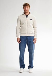 Bílá texturovaná bunda na zip s dlouhými rukávy, kombinovaná s modrými džínami a bílými teniskami. Bunda má na hrudi malý logo.