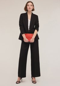 Zwarte op maat gemaakte blazer met geknipte revers, gecombineerd met wijde zwarte broek. Een rode clutch wordt voor zich gehouden; gespen met zwarte hakken maken de look af.