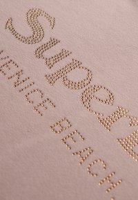 Tessuto beige con borchie metalliche dorate in rilievo che formano il testo "SUPPORT VENICE BEACH" con una superficie testurizzata.