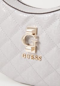 Betrukket lys grå håndtaske med et tekstureret logo-mønster og guld-tone "G" lukning over "GUESS" bogstaverne i midten.