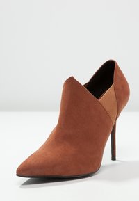 Stivale alla caviglia in suede marrone con tacco alto e punta affusolata, dotato di un design con ritagli e pannello elastico laterale per una facile vestibilità.
