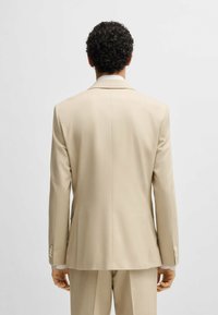 Giacca beige con vestibilità su misura, rever a punta e due spacchi posteriori. Il tessuto ha una texture liscia con una leggera lucentezza.