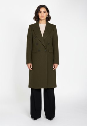 Manteau double boutonnage vert olive avec de grands boutons noirs, revers crantés, poches latérales et coupe droite. Associé à un pantalon noir à jambes larges.