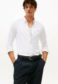 Camicia bianca con bottoni e maniche lunghe, abbinata a pantaloni blu navy e una cintura nera intrecciata. Logo piccolo e distintivo sulla camicia.