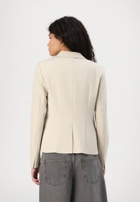 Beige-muotoiltu bleiseri, jossa takana on yksi nappi, slim fit -malli, pitkät hihat ja jämäkät hartiat, yhdistettynä harmaisiin leveälahkeisiin housuihin.