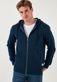 Sudadera con cremallera en color navy, con bolsillos frontales y capucha con cordones, fabricada en un tejido suave y texturizado. Camiseta blanca debajo, combinada con jeans azules.