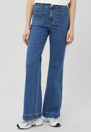 Femme portant un jean bleu taille haute à jambes larges avec des poches plaquées à l'avant et des baskets blanches, debout sur un fond clair uni.