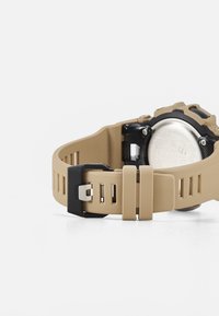  Bracelet de montre en silicone beige avec des accessoires en noir, présentant une surface texturée et des ouvertures pour la respirabilité, fixé à un boîtier en métal.