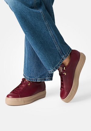 Baskets en cuir bordeaux avec une surface texturée, dotées de semelles en caoutchouc beiges et d'œillets dorés, assorties à un jean bleu à jambes larges.