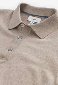 Beige Poloshirt aus strukturiertem Stoff, mit klassischem Kragen, drei grauen Knöpfen und einem gestickten Etikett mit dem Markennamen "NEXT" innen.