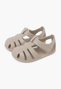 Sandalias de goma beige con diseño de puntera cerrada, que cuentan con correas horizontales y verticales. Correa ajustable en el tobillo con un acento de logo circular.