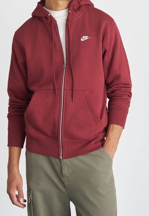 Homme portant un sweat à capuche zippé Nike bordeaux sur une chemise blanche et un pantalon vert olive, une main dans la poche, sur un fond uni.