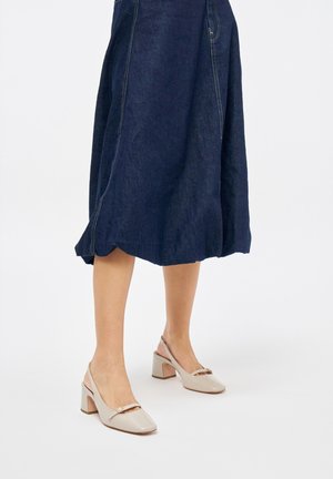 Tacchi slingback in patent beige con punta quadrata, tacco a blocco basso e dettagli decorativi sulla striscia. Abbinati a una gonna di denim scuro.