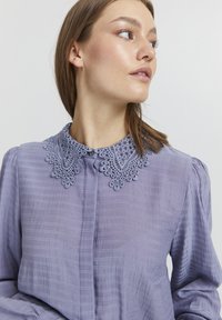 Blusa lavanda in tessuto texturizzato, con collare in pizzo dettagliato e chiusura a bottoni. Maniche lunghe con leggero volume alle spalle.