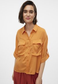 Camicia a maniche corte, con bottoni, di un vivace arancione con due tasche frontali e una vestibilità comoda. Tessuto liscio; presenta delle linguette per arrotolare le maniche.