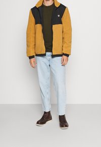 Żółto-czarna bluza z polaru na zamek błyskawiczny o teksturowanym wyglądzie, w połączeniu z zieloną koszulą, jasnymi dżinsami i brązowymi sznurowanymi butami.