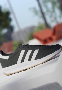Zapatilla Adidas en blanco y negro con tres rayas blancas y suela de goma apoyada sobre una superficie blanca, fondo exterior desenfocado.
