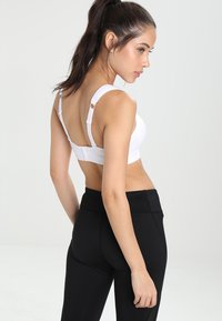 Sujetador deportivo blanco con tirantes ajustables y cierre de gancho. Combinado con leggings negros de cintura alta. Textura suave y elementos de diseño mínimos.
