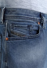 Vaqueros de mezclilla azules con un pequeño bolsillo frontal, botón de color cobre y un ligero desgastado. Diseño casual con detalles de costura visibles.