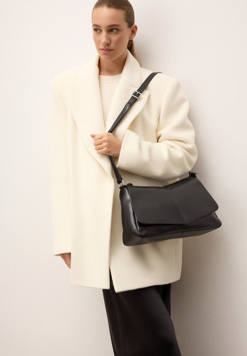 Borsa a tracolla in pelle nera con chiusura a pattina e tracolla regolabile, abbinata a un cappotto in misto lana color crema e a un maglione beige.