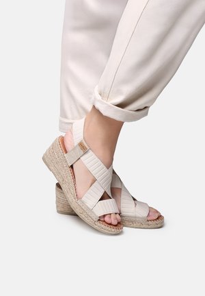 Feet wearing beige crisscross elastic strap wedge sandals paired with rolled-up light-colored pants against a plain background.