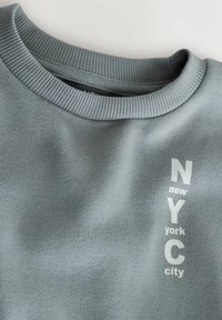 Sweatshirt gri deschis cu guler rotund, având inscripționat textul "NYC" și "new york city" în alb. Material moale cu o textură netedă.
