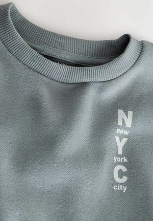 Light gray sweatshirt with a crew neck, featuring printed text "NYC" and "new york city" in white. Soft fabric with a smooth texture.