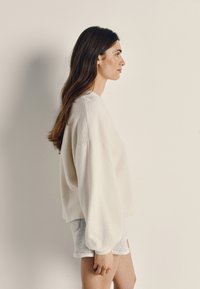 Fleecinis megztinis smėlio spalvos su tekstūruota paviršiaus struktūra, laisvo silueto, su nukritusiomis pečių linijomis ir ribinėmis rankogaliais. Derinamas su pilkais šortais.