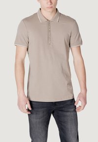 Polo beige con patta anteriore, maniche corte e rifiniture bianche. Presenta un logo al centro. Abbinato a jeans scuri.