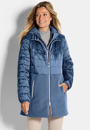 Frau trägt einen blau gesteppte und Fleece-Mantel mittlerer Länge mit Kapuze, steht und lächelt, Hände in den Taschen, beige Hose sichtbar.