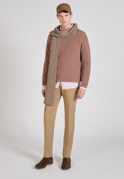 Uomo che indossa un berretto marrone, un maglione marrone a coste, una camicia a righe, una lunga sciarpa beige lavorata a maglia, pantaloni beige e stivali in camoscio marrone, in piedi su uno sfondo chiaro.