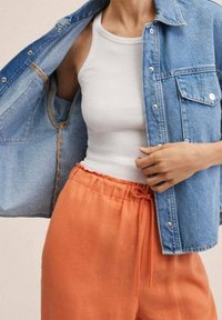 Denim jacket with blue wash, featuring metal snaps and pockets. Underneath, a white tank top and orange elastic-waist pants with drawstrings.