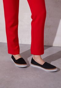 Pieds portant des chaussures noires à enfiler avec des semelles marron et blanche, associés à un pantalon droit rouge vif sur un sol gris.