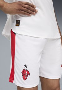 Shorts de sport blancs avec des panneaux latéraux rouges. Présentent un logo noir d'une fox stylisée et une petite étoile noire au-dessus. Tissu texturé.