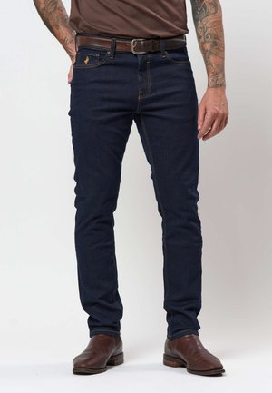 Mężczyzna ubrany w ciemnoniebieskie dżinsy, brązowy skórzany pasek, brązowe buty i brązową koszulę, z widocznymi tatuażami na ramionach od łokcia w dół.