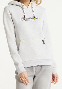 Žena v světle šedé mikině s kapucí, černými a žlutými šňůrkami a přední kapsou, v kombinaci se světle šedými džíny.