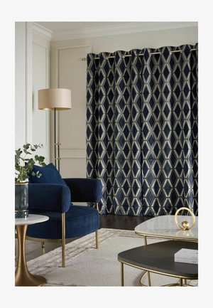 Blauer Samtsessel mit goldfarbenen Beinen, Vorhänge mit geometrischem Muster, Stehlampe, kleine runde Tische mit goldenen Akzenten und eine Pflanze in einer Vase.