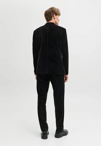 Juodas aksominis kostiumas su atverčiama atlapo tipo švarku ir lieknų šoninių siūlių kelnėmis, pasižyminčiomis švelnia tekstūra ir subtiliu blizgesiu, derinamas su juodais batais.