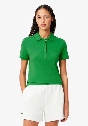 Polo vert avec col et manches courtes, présentant une patte de boutonnage. Fabriqué en tissu texturé, associé à un short blanc.
