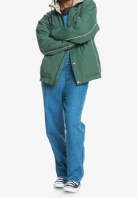 Zielona kurtka puchowa z beżowym kołnierzem i białymi lamówkami, zestawiona z jasnoniebieskimi luźnymi dżinsami i czarno-białymi trampkami.