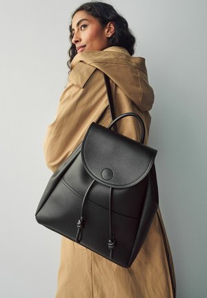 Schwarzer Leder-Rucksack mit einer abgerundeten Klappe, Kordelzugverschluss und subtilen Stickdetails, getragen über einem beigefarbenen Mantel.