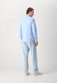 Camicia azzurra a maniche lunghe con colletto button-down, indossata con pantaloni in denim chiaro e sneakers bianche, vista da dietro.