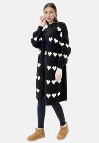 Zwarte gebreide cardigan met witte hartpatronen, open voorkant en lange mouwen. Draagt met zwarte legging en bruine enkellaarsjes.